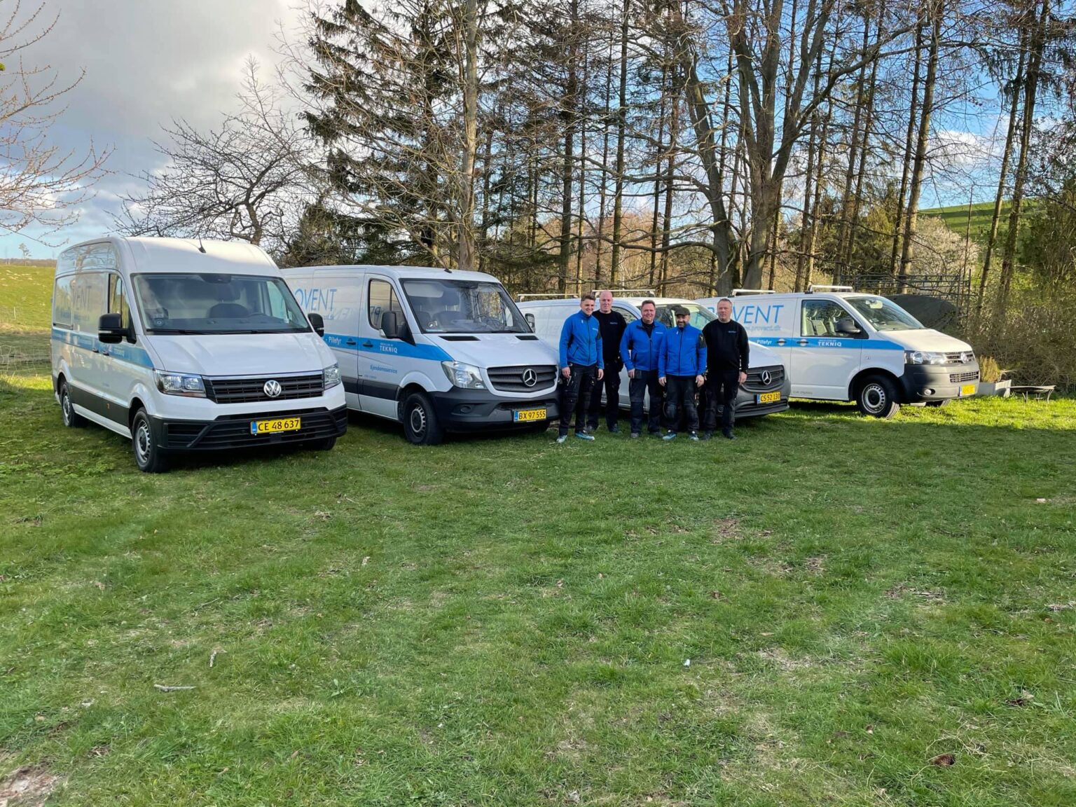 Fem mænd står foran servicebiler i naturen.