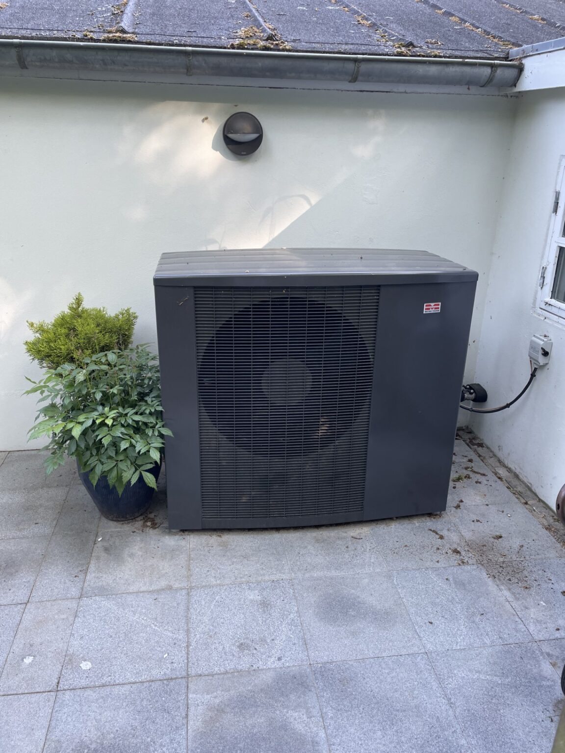 Udendørs varmepumpe ved siden af grøn plante.